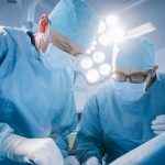 Low Angle Shot In Operating Room of Two Surgeons During the Surgery Procedure Bending Over Patient with Instruments. Professional Doctors in Modern Hospital