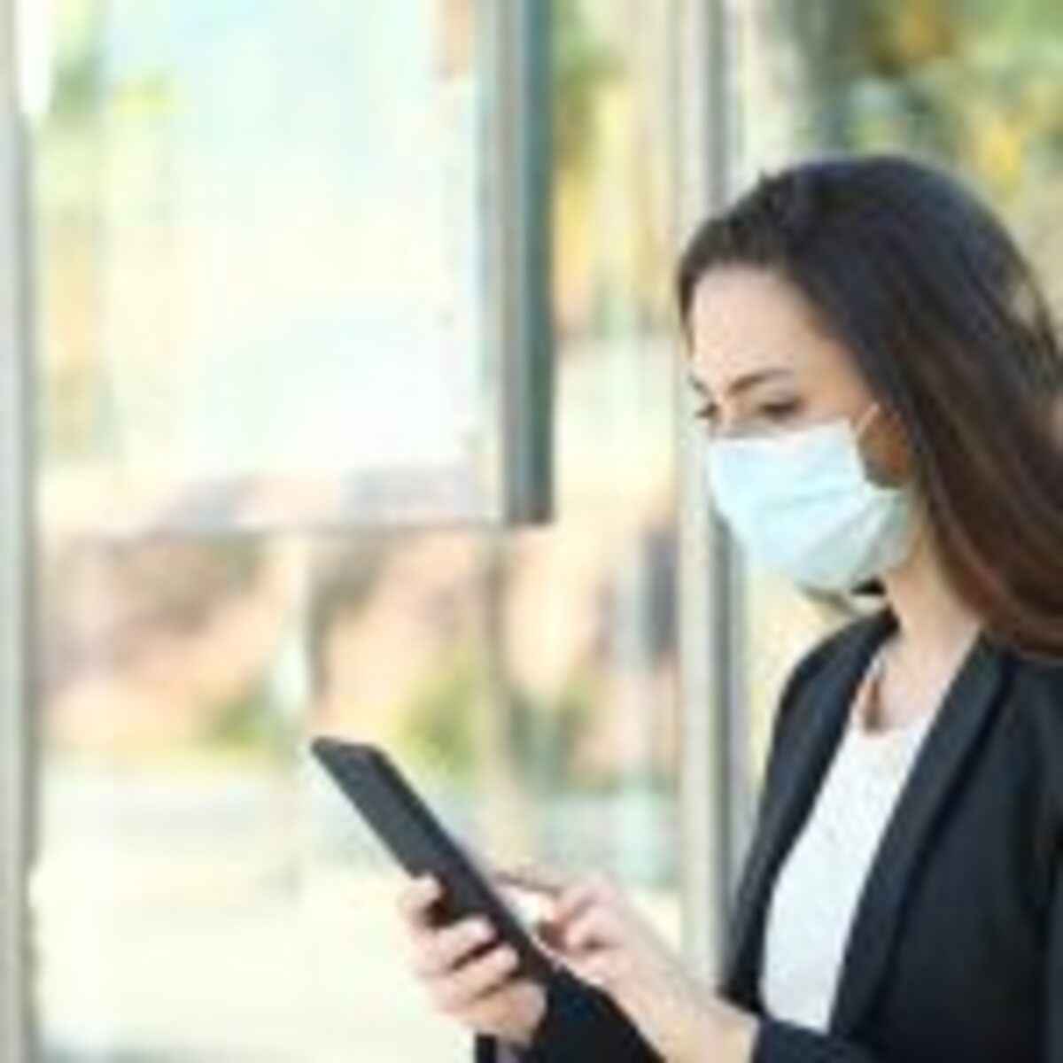 Woman wearing a protective mask using mobile phone sitting in a bus stop