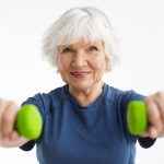 Active energetic happy elderly Caucasian female with gray hair enjoying physical exercises indoors, training at home using dumbbells, smiling broadly at camera. Selective focus on woman’s face