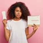 Photo of discontent lovely curly haired woman looks at calendar with marked pms days, holds sanitary napkin for hygiene protection, has bad mood during menstruation, wears casual white t shirt