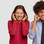 Photo of stressful girls clench teeth, plug ears, ignore unpleasant sound, stand closely, dressed in casual outfit, isolated over white backgrounf with free space for your advertisement or promotion