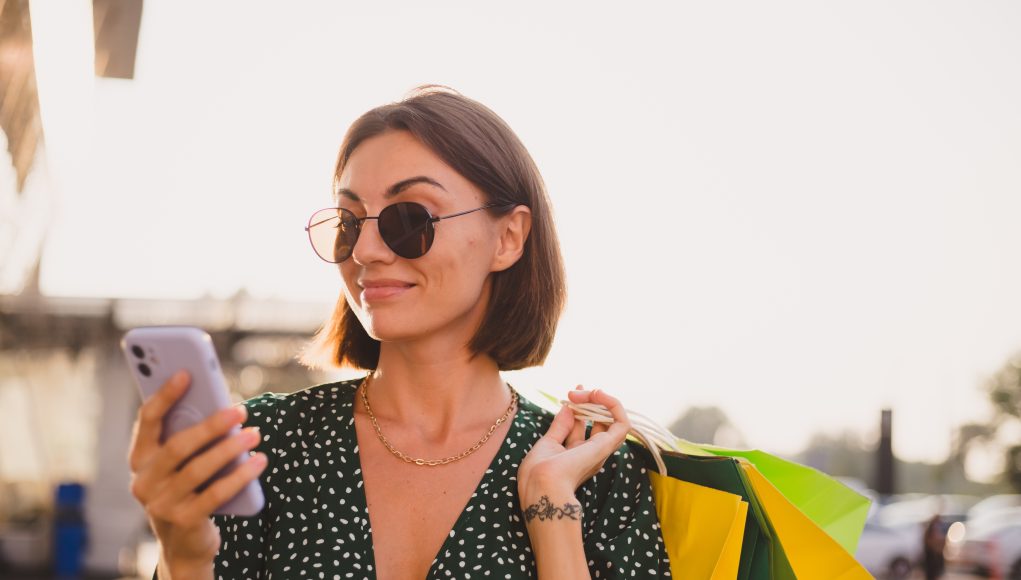 Woman at sunset with colorful shopping bags and parking lot by shopping mall happy with mobile phone