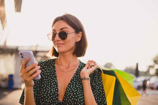 Woman at sunset with colorful shopping bags and parking lot by shopping mall happy with mobile phone