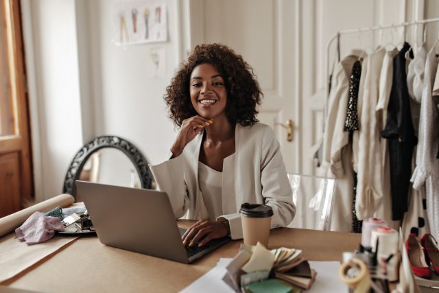 Charming young dark-skinned woman in stylish jacket and blouse smiles, looks at camera, works in la