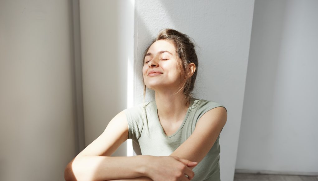 Portrait of tender young beautiful girl smiling with closed eyes enjoying morning sunlights sitting on floor over white wall.