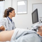 Careful female doctor in white coat sitting in front of an ultrasound apparatus and conducting abdominal diagnostics with transducer
